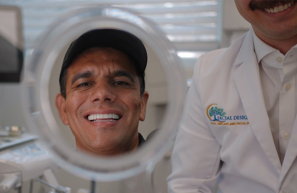 A man smiles while looking at his reflection in a circular mirror inside a dental office, standing beside a clinician wearing a white coat embroidered with the Facial Designs practice logo.