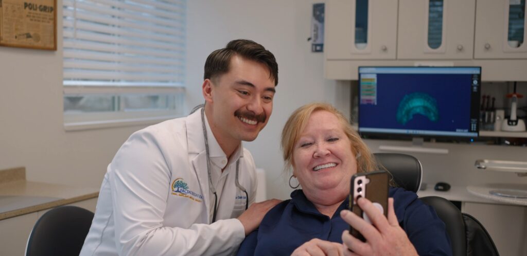 Dr. Zane Clark-Gold in a white coat embroidered smiles alongside a patient seated in a dental chair as they look at a smartphone together, with a 3D digital dental model displayed on a nearby monitor. showing you why choose a prosthodontist