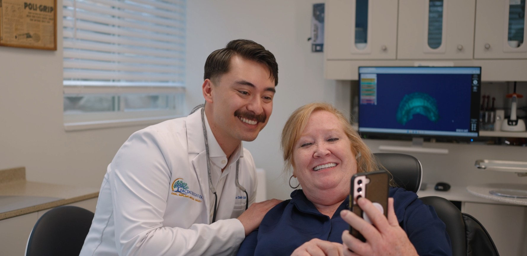 Dr. Zane Clark-Gold in a white coat embroidered smiles alongside a patient seated in a dental chair as they look at a smartphone together, with a 3D digital dental model displayed on a nearby monitor. showing you why choose a prosthodontist