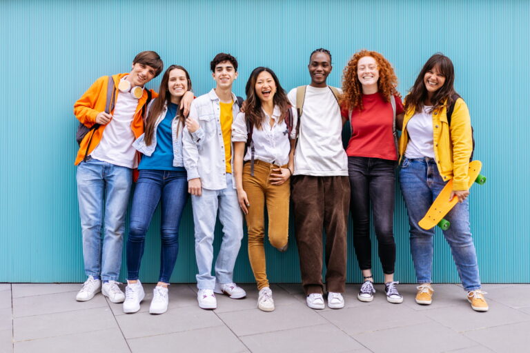 Portrait of seven young student friends leaning on blue wall. Diverse teenage classmates looking at camera standing.