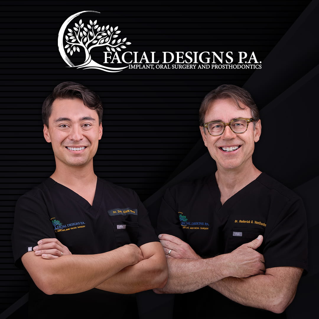 Dr. Clark-Gold (left) and Dr. VanSurksum (right) wearing black medical scrubs stand side by side against a dark background. Both have their arms crossed and are smiling. Their uniforms display embroidered names along with “Facial Designs P.A. Implant, Oral Surgery and Prosthodontics.” Above them is a circular tree‑themed logo with the same practice name and tagline.