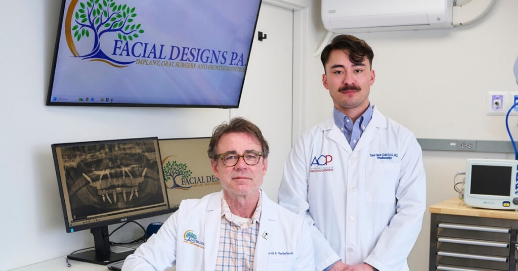 Dr. VanSurksum and Dr. Clark-Gold in white lab coats pose in a treatment room. One sits on a stool while the other stands beside them. Behind them, two computer monitors display a dental X‑ray and a logo reading “Facial Designs PA Implant, Oral Surgery and Prosthodontics,” featuring a stylized tree with green leaves. The room includes medical equipment, drawers, and an air conditioning unit.