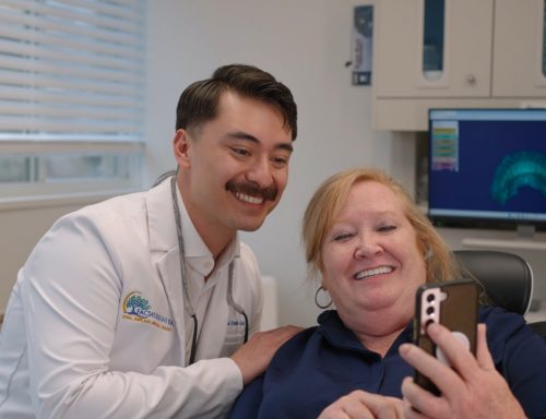 Dr. Zane Clark-Gold in a white coat embroidered smiles alongside a patient seated in a dental chair as they look at a smartphone together, with a 3D digital dental model displayed on a nearby monitor.