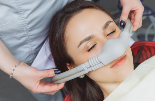 A person reclines with eyes closed while wearing a nitrous oxide nasal mask, as a healthcare professional adjusts the mask in a clinical setting.