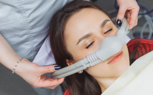 A person reclines with eyes closed while wearing a nitrous oxide nasal mask, as a healthcare professional adjusts the mask in a clinical setting.