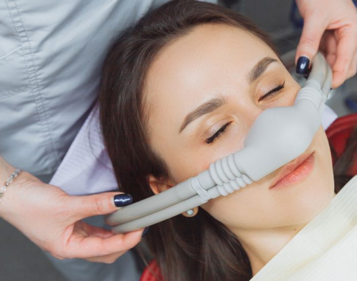 A person reclines with eyes closed while wearing a nitrous oxide nasal mask, as a healthcare professional adjusts the mask in a clinical setting.