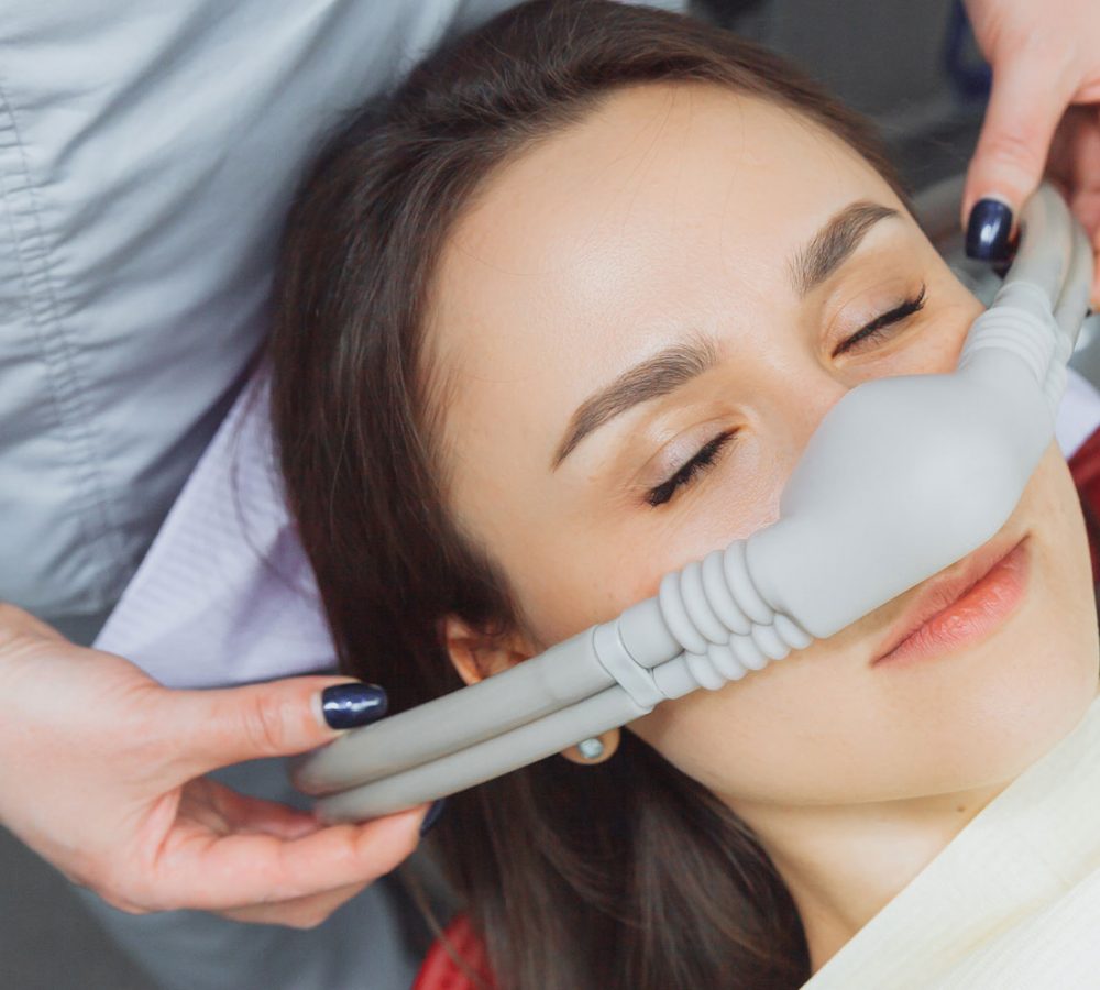 A person reclines with eyes closed while wearing a nitrous oxide nasal mask, as a healthcare professional adjusts the mask in a clinical setting.